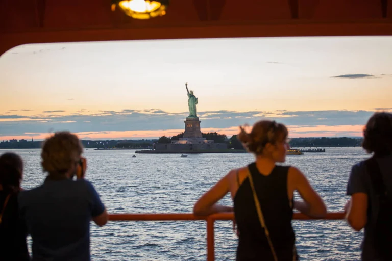 Silhouetten van reizigers op een veerboot kijken uit over het water naar het Vrijheidsbeeld in New York tijdens zonsondergang