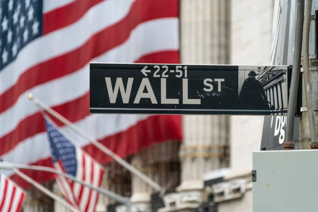 Close-up van een zwart straatbord met de tekst Wall Street, met wapperende Amerikaanse vlaggen en een zuilengevel op de achtergrond