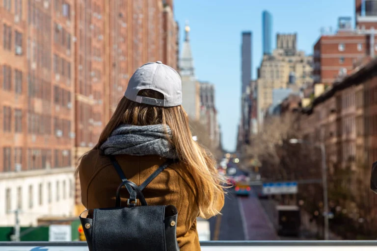 Vrouw met rugzak en pet kijkt van bovenaf vanaf de High Line naar een drukke straat met wolkenkrabbers op de achtergrond