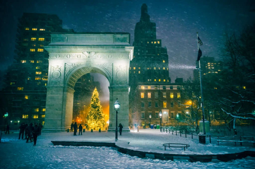 Verlichte Washington Square boog in New York tijdens sneeuwval met een fonkelende kerstboom en wandelaars in de avond