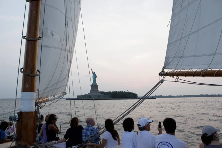Toeristen op een zeilboot met witte zeilen kijken uit over het water naar het Vrijheidsbeeld in New York bij zonsondergang