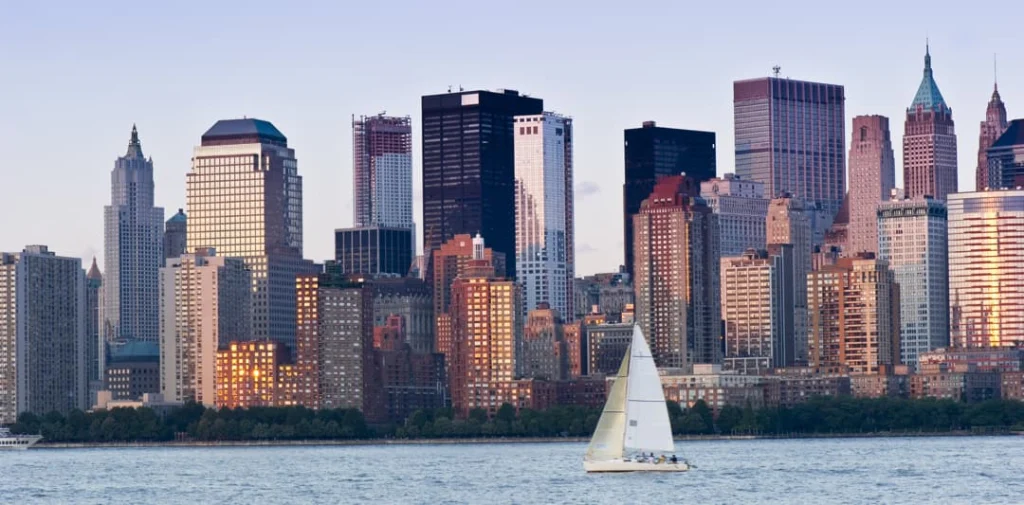 Een zeilboot vaart over het water terwijl de wolkenkrabbers van Lower Manhattan oplichten in het avondlicht