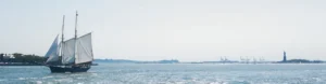 Een historisch zeilschip met witte zeilen vaart over het water in de haven van New York, met op de achtergrond het Vrijheidsbeeld en havenkranen