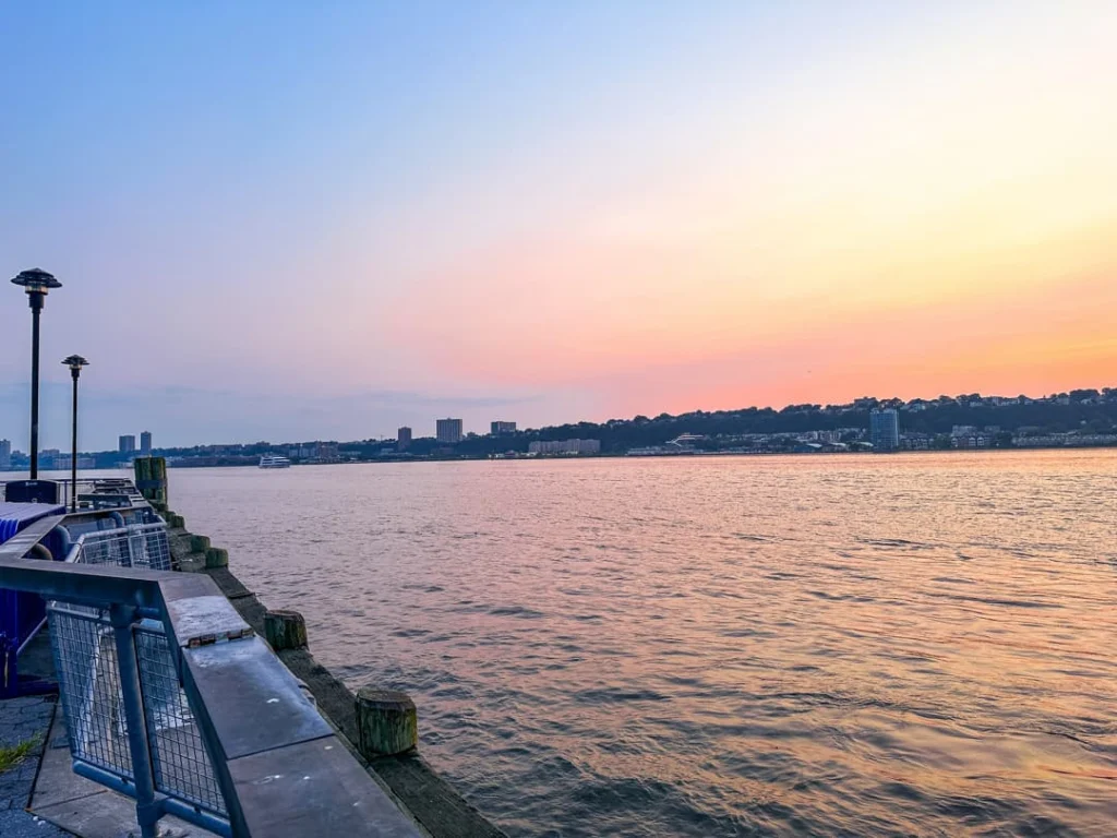 Pastelkleurige zonsondergang boven de Hudson rivier met een verlichte pier op de voorgrond en de skyline vaag zichtbaar aan de overkant
