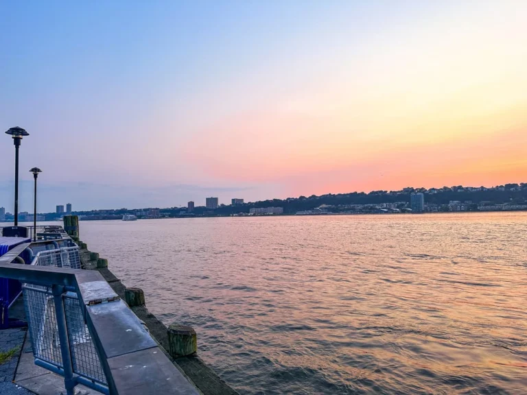 Pastelkleurige zonsondergang boven de Hudson rivier met een verlichte pier op de voorgrond en de skyline vaag zichtbaar aan de overkant