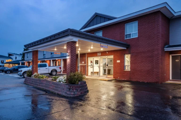 Bakstenen hotelgebouw met overkapte ingang en verlichte ramen op een natte parkeerplaats bij regenachtige schemering