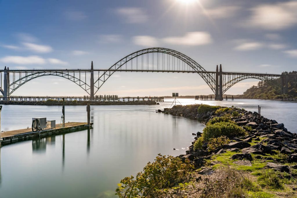Grote stalen boogbrug over de baai van Newport met een steiger en rotsachtige kade op de voorgrond onder een felle zon