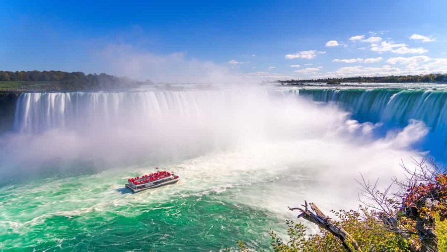 Een rode toeristenboot vaart door opspattend water voor de machtige Niagara watervallen onder een blauwe hemel