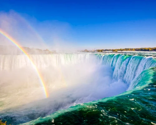 Brede waterval van de Niagara Falls met opspattend water, mist en een dubbele regenboog boven de rivier