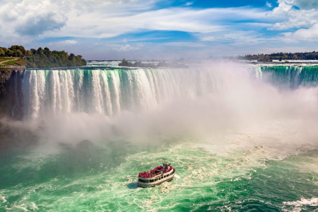 Een rondvaartboot vol toeristen vaart door opspattend water voor de machtige Niagara Falls onder een blauwe lucht