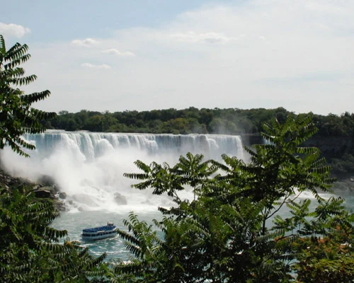 Een wit toeristenbootje vaart door opspattend water naar de brede Niagara Falls, met bomen op de voorgrond