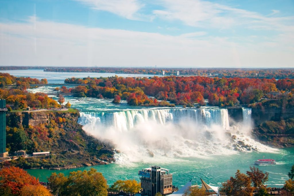 Luchtfoto van de bruisende Niagara Falls omringd door bomen met herfstkleuren en een boottochtje op het turquoise water
