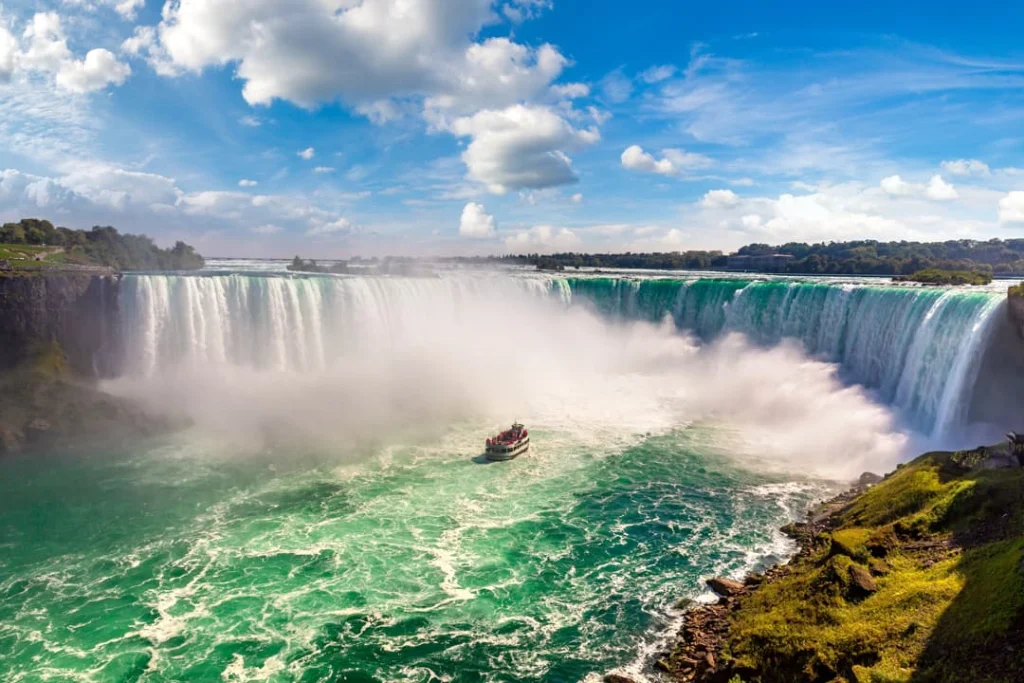 Een toeristenboot vaart door opspattende nevel vlak bij de krachtige hoefijzervormige Niagara watervallen onder een blauwe wolkenlucht
