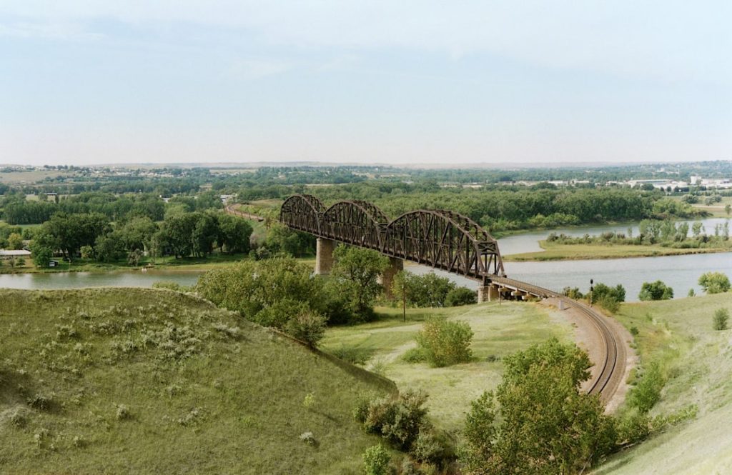 Uitzicht vanaf een grasheuvel op een lange ijzeren spoorbrug met bogen die over een rivier met spoorrails naar de horizon loopt