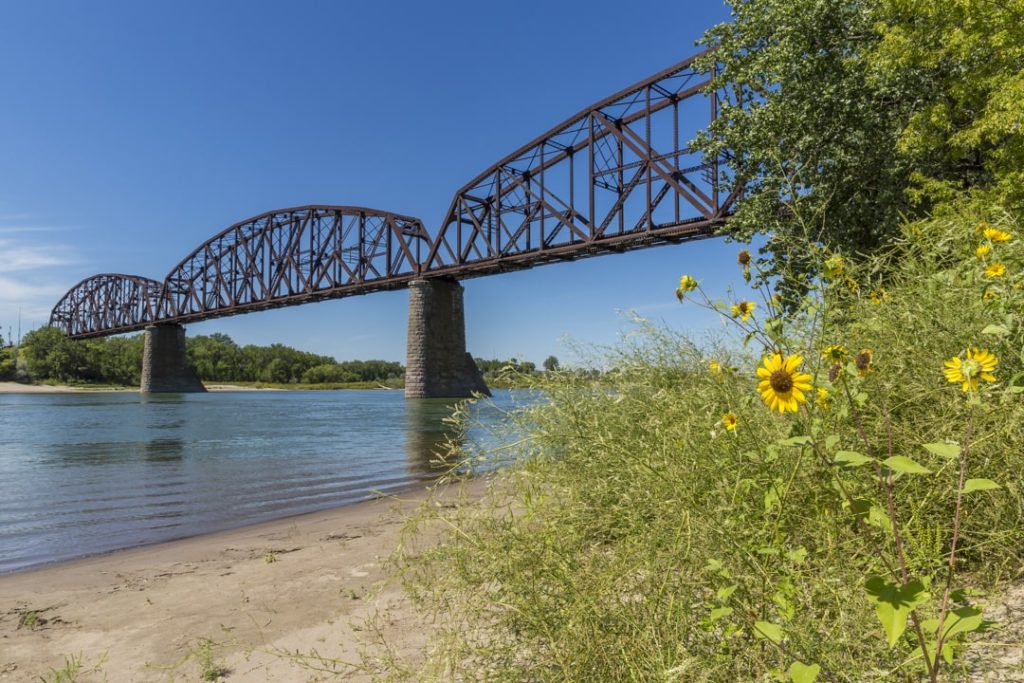 Roestbruine ijzeren spoorbrug met bogen over een brede rivier, met gele zonnebloemen en struiken op de oever op de voorgrond