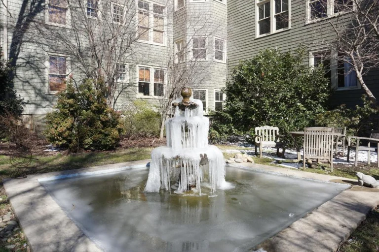 Een bevroren waterfontein met hangende ijspegels staat in een besneeuwde tuin tussen houten bankjes en gebouwen in North Carolina