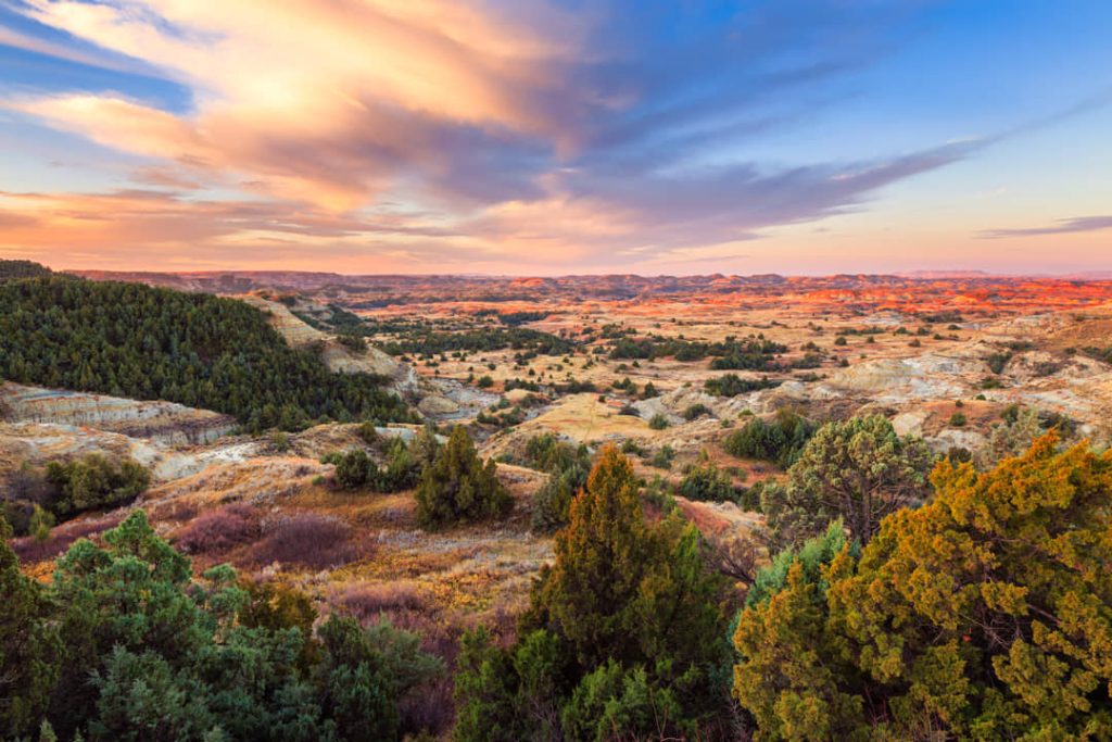 Glooiend landschap van geërodeerde heuvels en jeneverbesbomen in warme kleuren tijdens zonsondergang in North Dakota