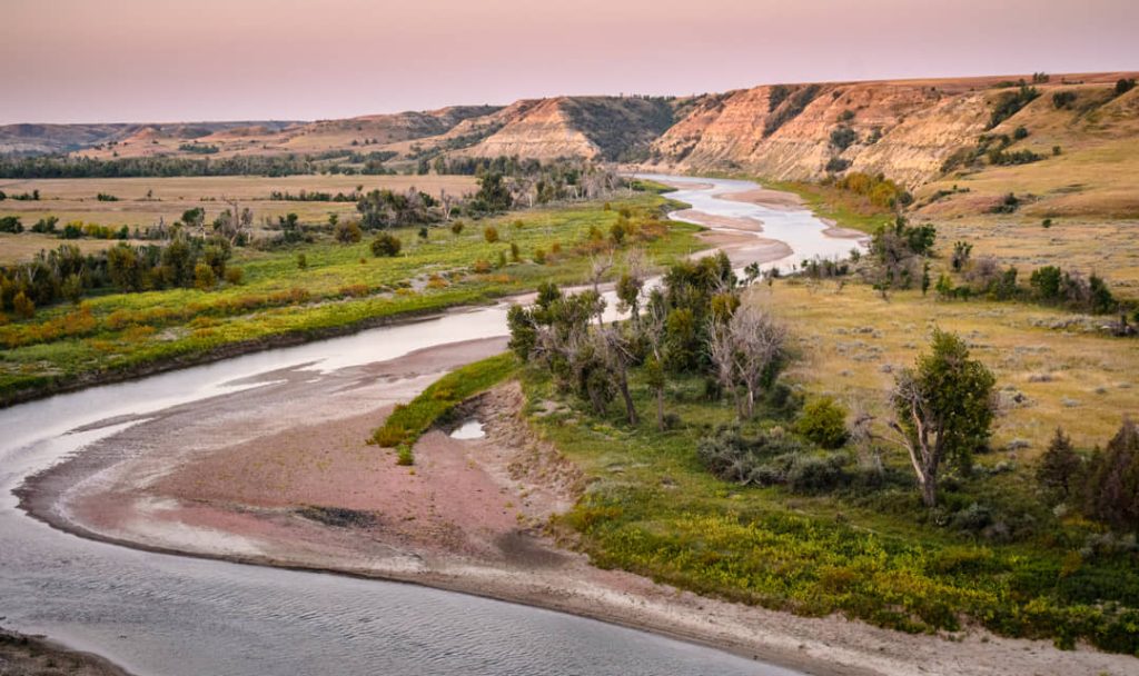 Luchtfoto van een kronkelende rivier tussen glooiende, deels beboste heuvels in de badlands van North Dakota bij ochtendlicht