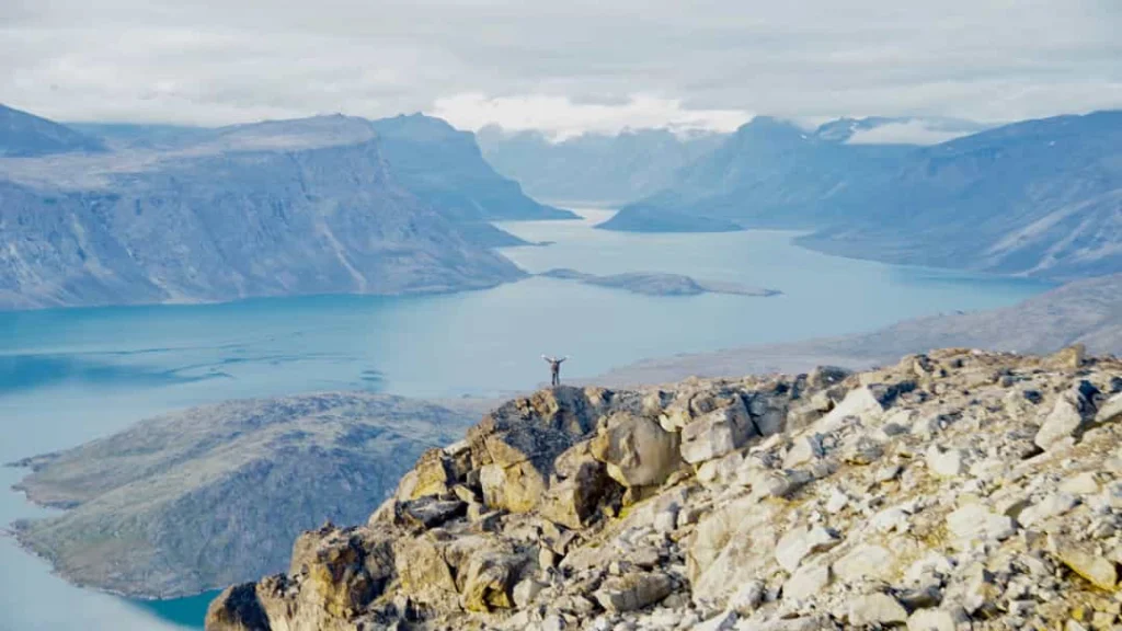 Wandelaar met gestrekte armen staat op een rotsachtige bergtop met uitzicht op de blauwe Pangnirtung Fjord in Nunavut