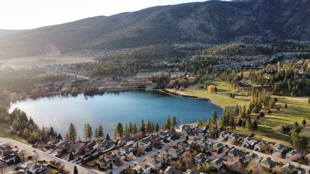 Luchtfoto van een klein blauw meer omringd door dennenbos, een golfterrein en huizenwijken tegen een heuvel in de Okanagan Valley