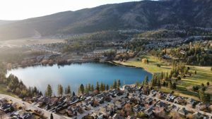 Luchtfoto van een klein blauw meer omringd door dennenbos, een golfterrein en huizenwijken tegen een heuvel in de Okanagan Valley