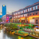 Verlicht kanaal in de wijk Bricktown met bakstenen pakhuizen, terrassen en een rondvaartboot bij avondlicht