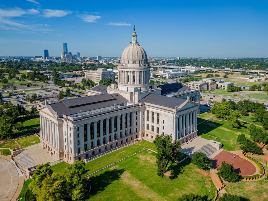 Zandkleurig koepelgebouw met zuilenrijen op de voorgrond, met de wolkenkrabbers van Oklahoma City zichtbaar aan de horizon