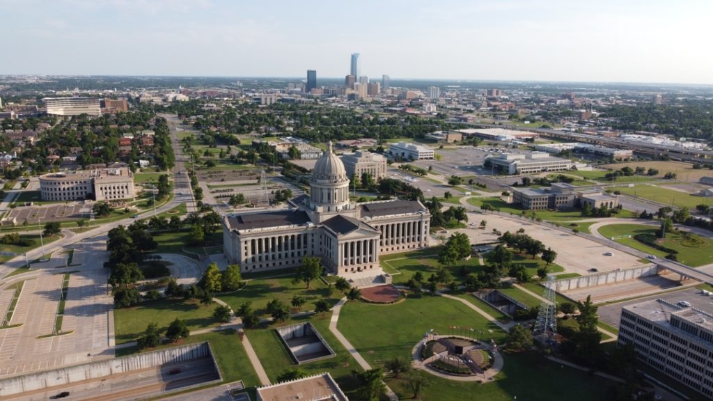 Luchtfoto van het koepelgebouw van het Oklahoma State Capitol omgeven door gazons en parkeerterreinen, met de wolkenkrabbers van Oklahoma City in de verte
