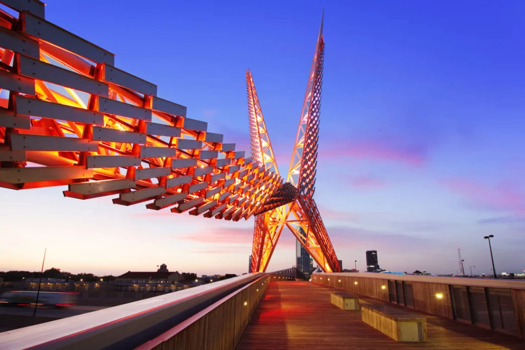 Verlichte voetgangersbrug met een opvallende metalen sculptuur in de vorm van vleugels tegen een roze avondlucht