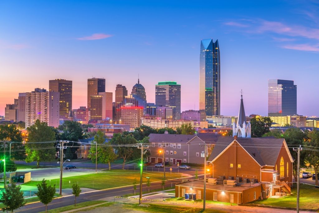 Verlichte wolkenkrabbers van Oklahoma City, waaronder een opvallende geknikte glazen toren, met een bakstenen kerk op de voorgrond in de schemering
