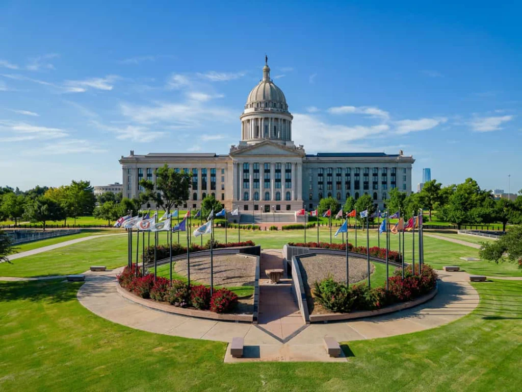 Voorgevel van het Oklahoma State Capitol met zuilen en koepel, met een rond plein met vlaggenmasten en bloemperken ervoor