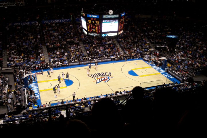 Basketbalveld met het logo van de Oklahoma City Thunder, gezien van bovenaf vanaf een hoge tribune vol publiek