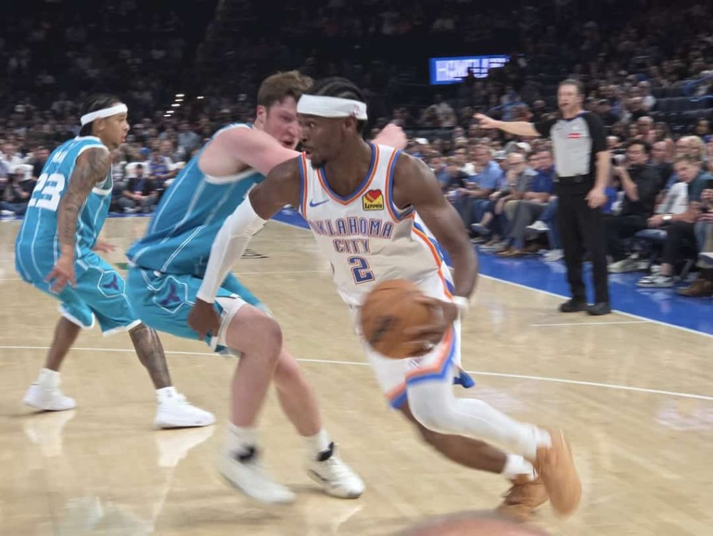 Basketbalspeler in het shirt van Oklahoma City dribbelt met de bal langs een verdediger tijdens een wedstrijd