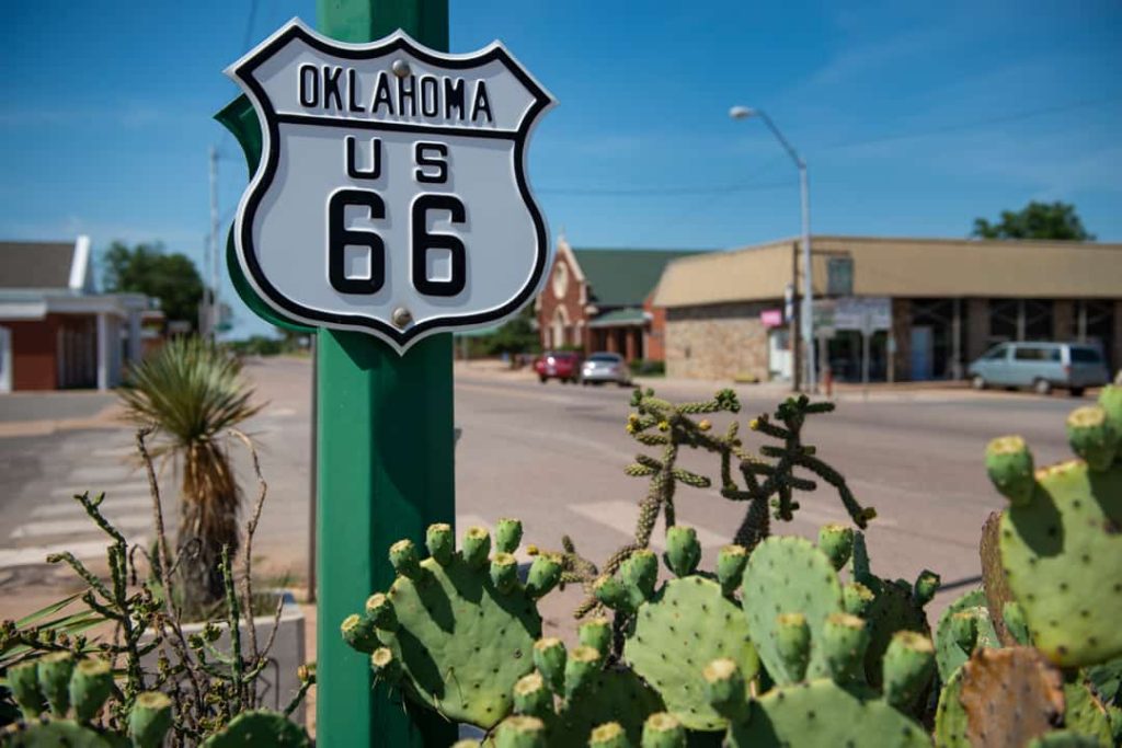 Wit schildvormig bord met de tekst oklahoma us 66 tussen cactussen langs een straat met winkels