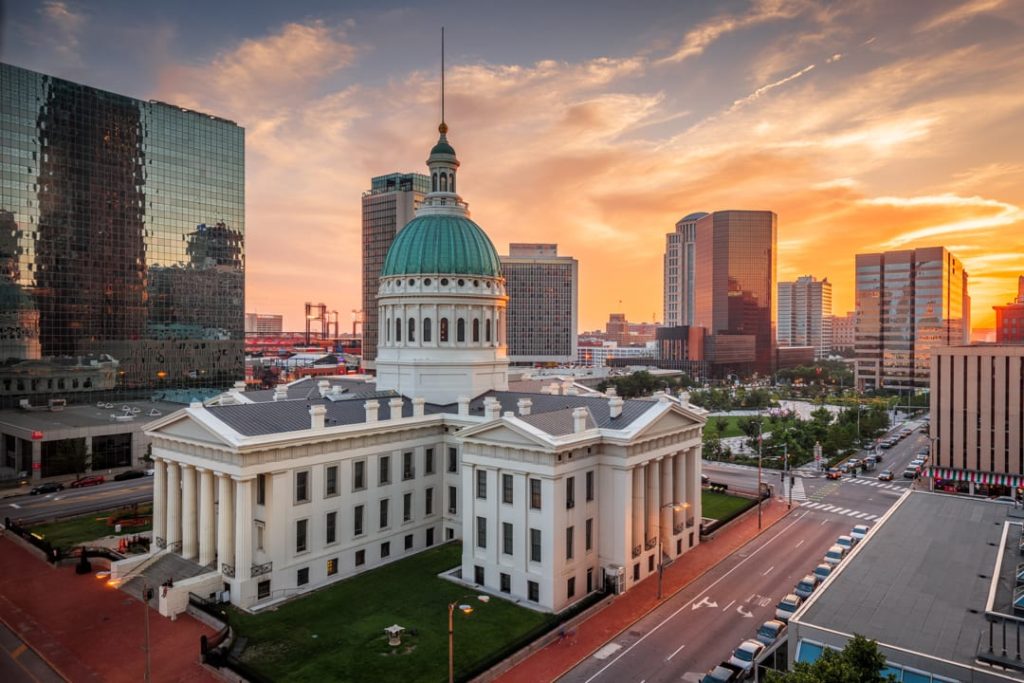 De groene koepel van de Old Courthouse in St. Louis afgetekend tegen een oranje avondlucht, tussen moderne wolkenkrabbers