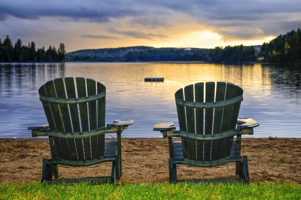 Twee lege houten Adirondack stoelen staan op een zandstrand met uitzicht op een meer tijdens een gouden zonsondergang