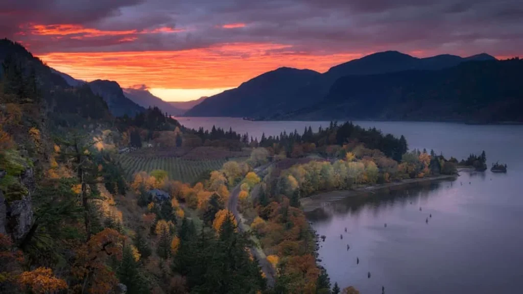 Vanaf een hoge uitkijk over de Columbia River Gorge in Oregon kleuren de wolken rood en oranje bij zonsondergang, met herfstbomen langs de rivieroever