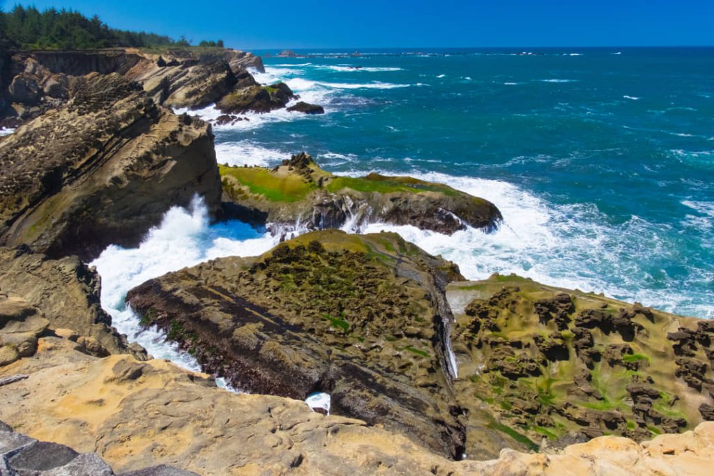 Schuimende golven slaan tegen begroeide rotsformaties langs de ruige kustlijn van Oregon onder een blauwe hemel