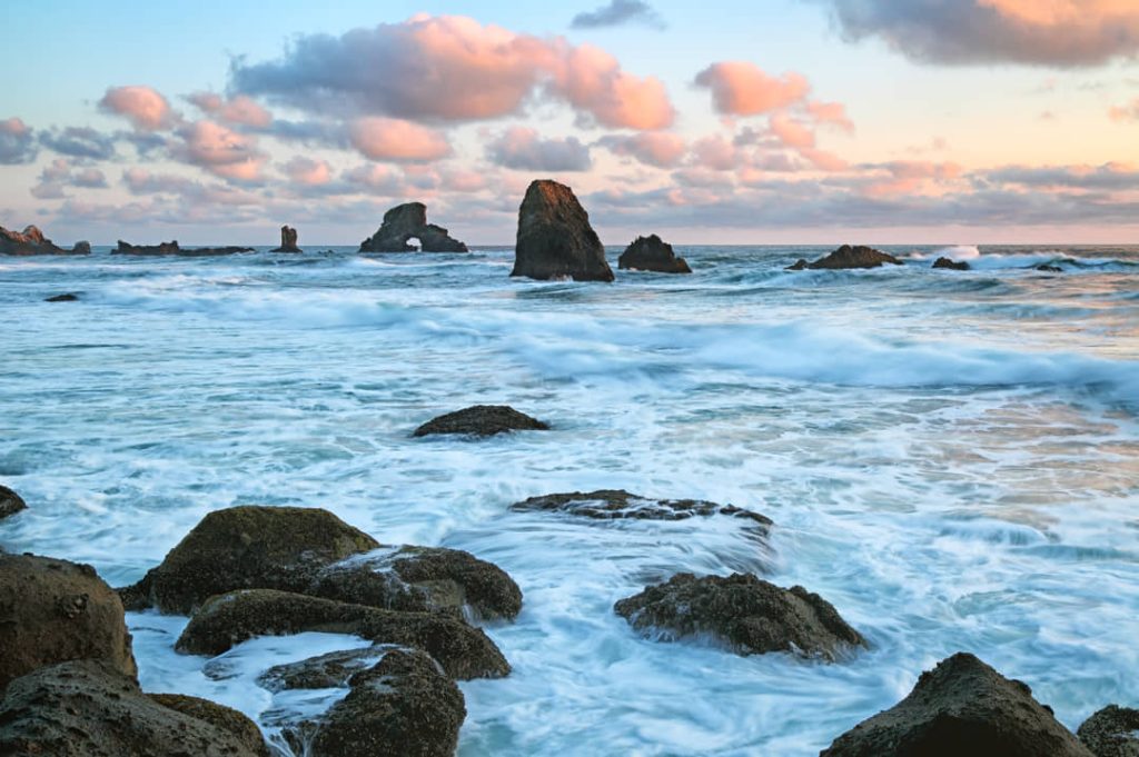 Ruige rotsen op de voorgrond met wazige zeestapels in de branding onder een roze bewolkte avondlucht aan de kust van Oregon