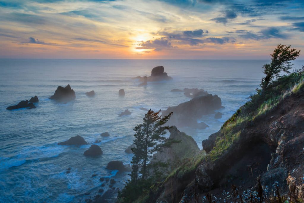 Ruige kustlijn met verspreide rotsformaties in zee, gehuld in mist, terwijl de zon ondergaat aan de kust van Oregon