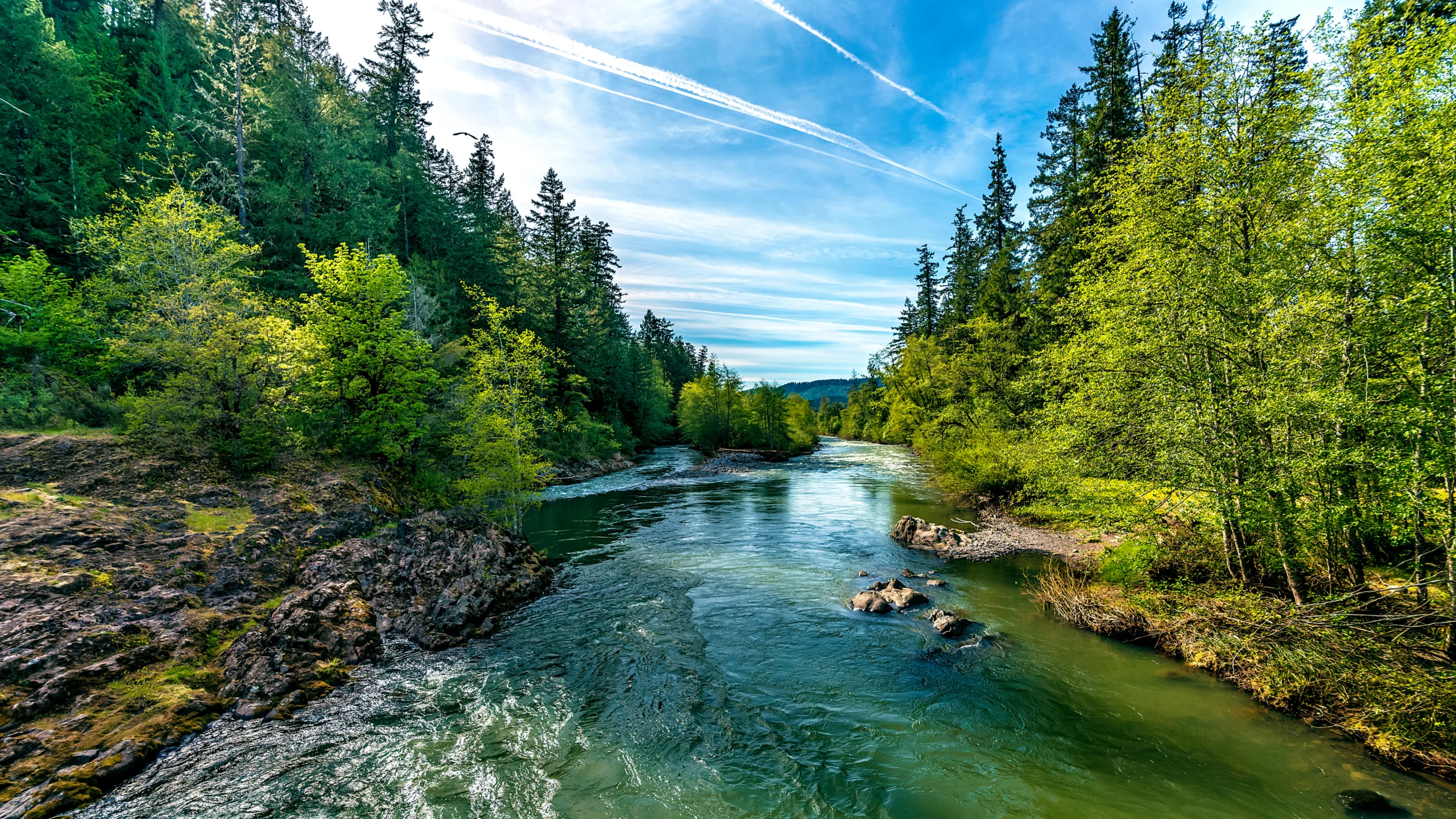 Een helder groenblauwe rivier stroomt tussen rotsen door een dicht naaldbos onder een blauwe lucht met wolkensporen in Oregon