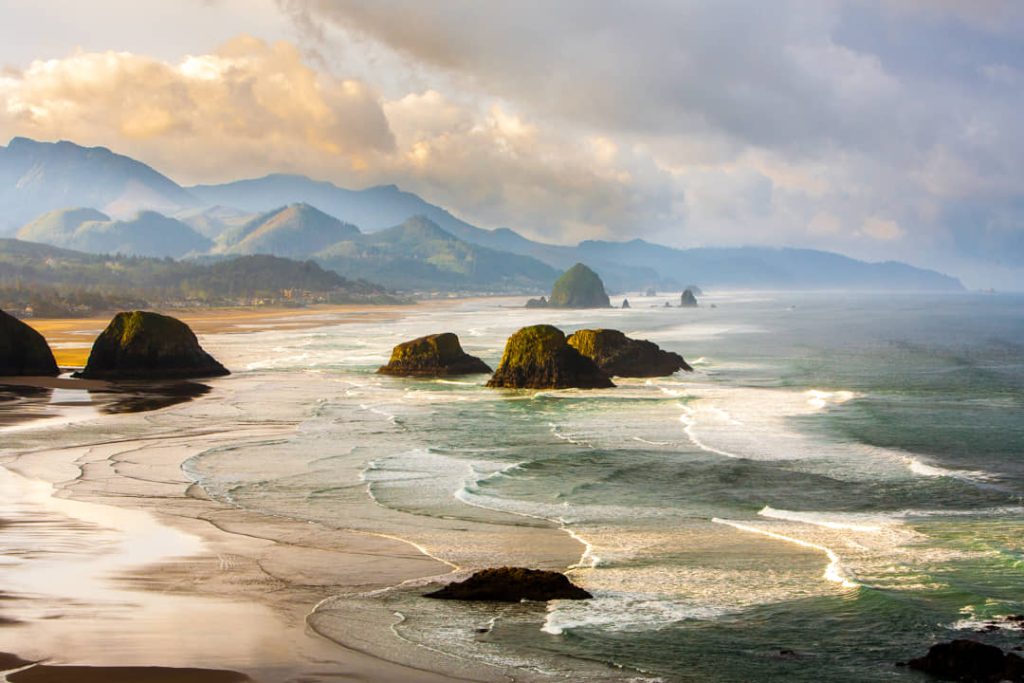 Verhoogd uitzicht over een breed zandstrand met verspreide zeestapels in de golven en beboste bergen gehuld in mist in Oregon