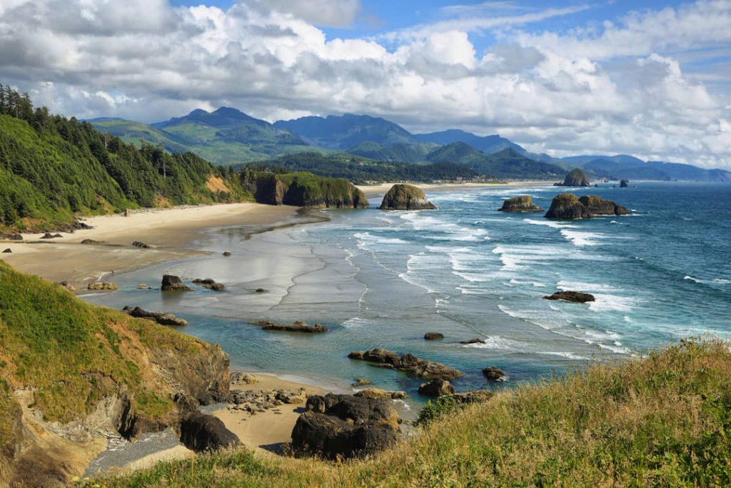 Panoramisch uitzicht vanaf een groene heuvel over Cannon Beach met rotsachtige zeestapels in het water en bergen op de achtergrond in Oregon