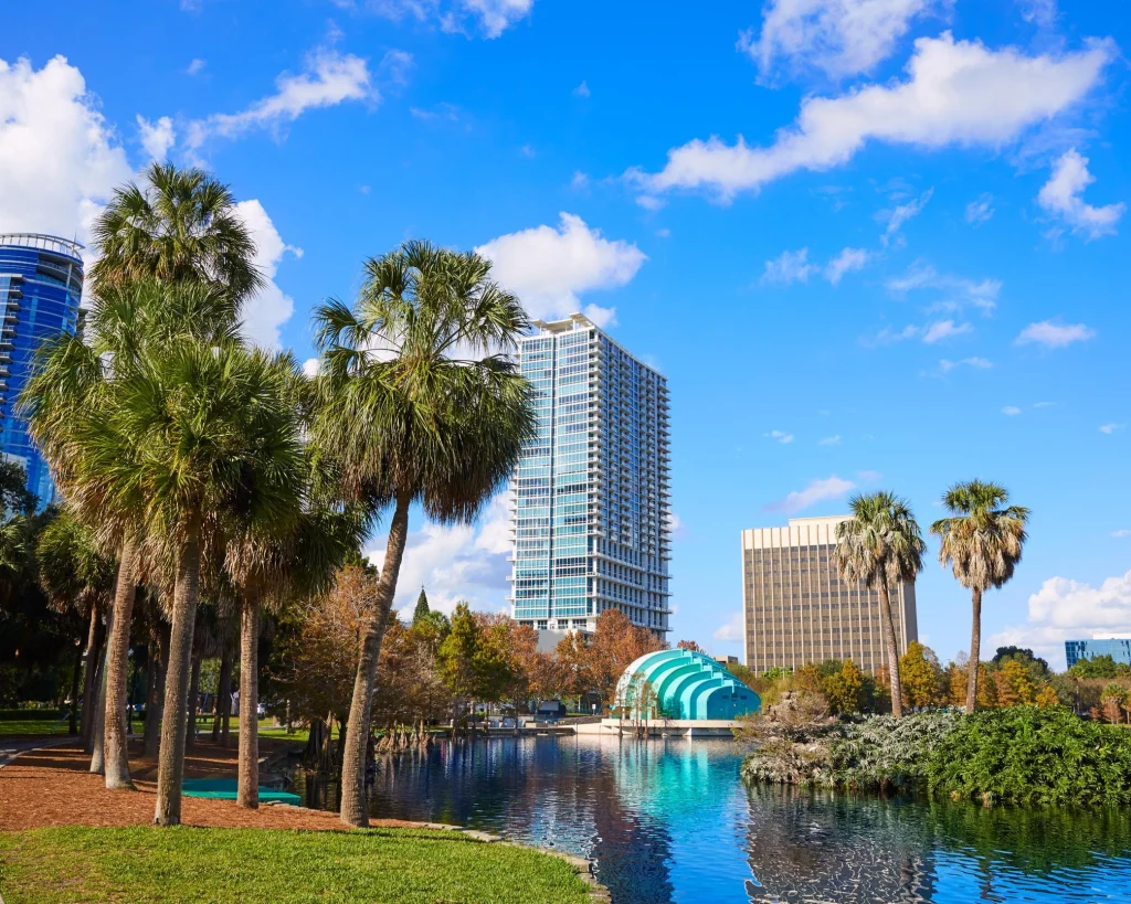 Palmbomen en groen gras rond het water van Lake Eola Park in Orlando met wolkenkrabbers en een koepelvormige muziekkoepel op de achtergrond
