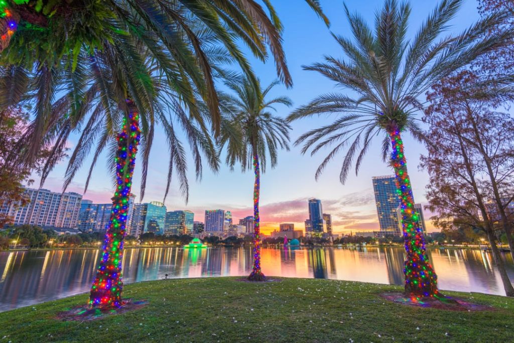 Drie palmbomen versierd met kleurrijke lichtjes bij het water van Lake Eola Park met de verlichte skyline van Orlando op de achtergrond