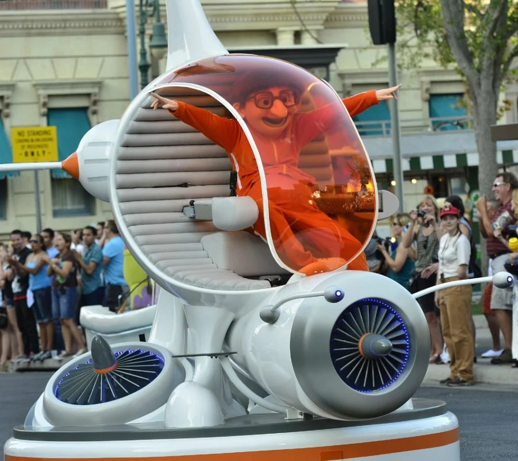 Een personage met opgeheven armen zit in een futuristisch parade voertuig terwijl bezoekers langs de kant toekijken