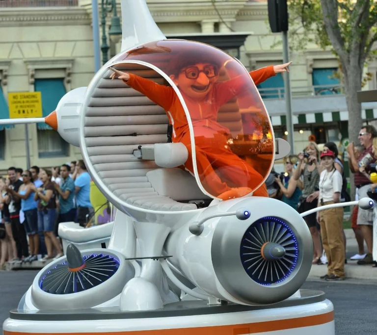 Een personage met opgeheven armen zit in een futuristisch parade voertuig terwijl bezoekers langs de kant toekijken