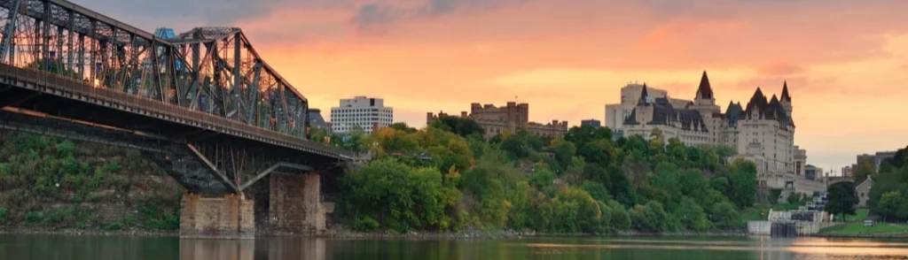 IJzeren spoorbrug over de rivier met op de achtergrond het kasteelachtige Château Laurier onder een oranje avondlucht in Ottawa