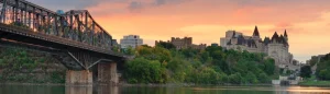 IJzeren spoorbrug over de rivier met op de achtergrond het kasteelachtige Château Laurier onder een oranje avondlucht in Ottawa