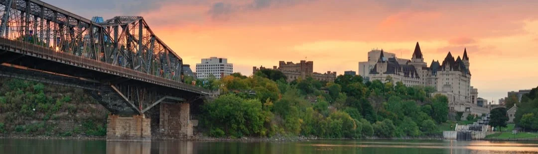 IJzeren spoorbrug over de rivier met op de achtergrond het kasteelachtige Château Laurier onder een oranje avondlucht in Ottawa